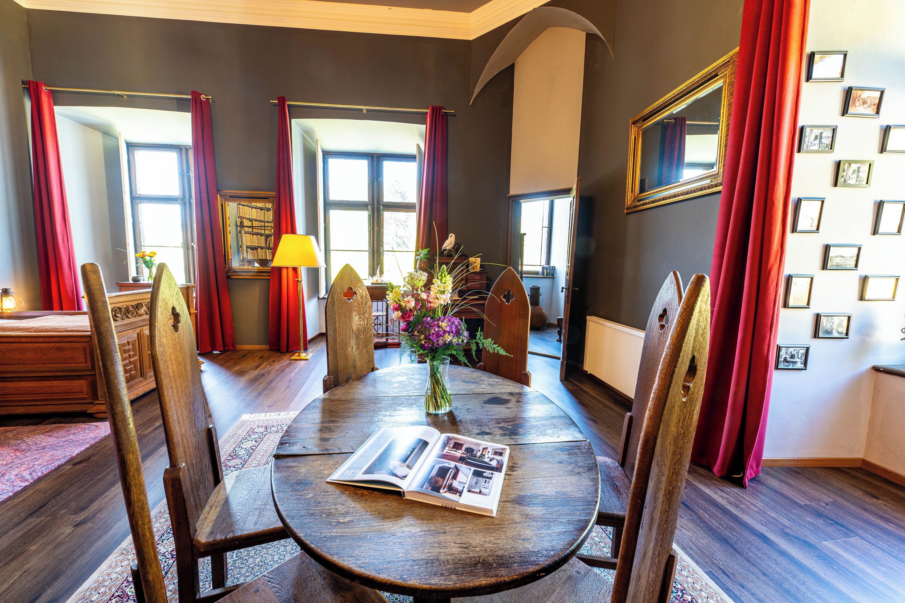 Un comedor con una mesa redonda de madera, cortinas rojas y un jarrón con flores.