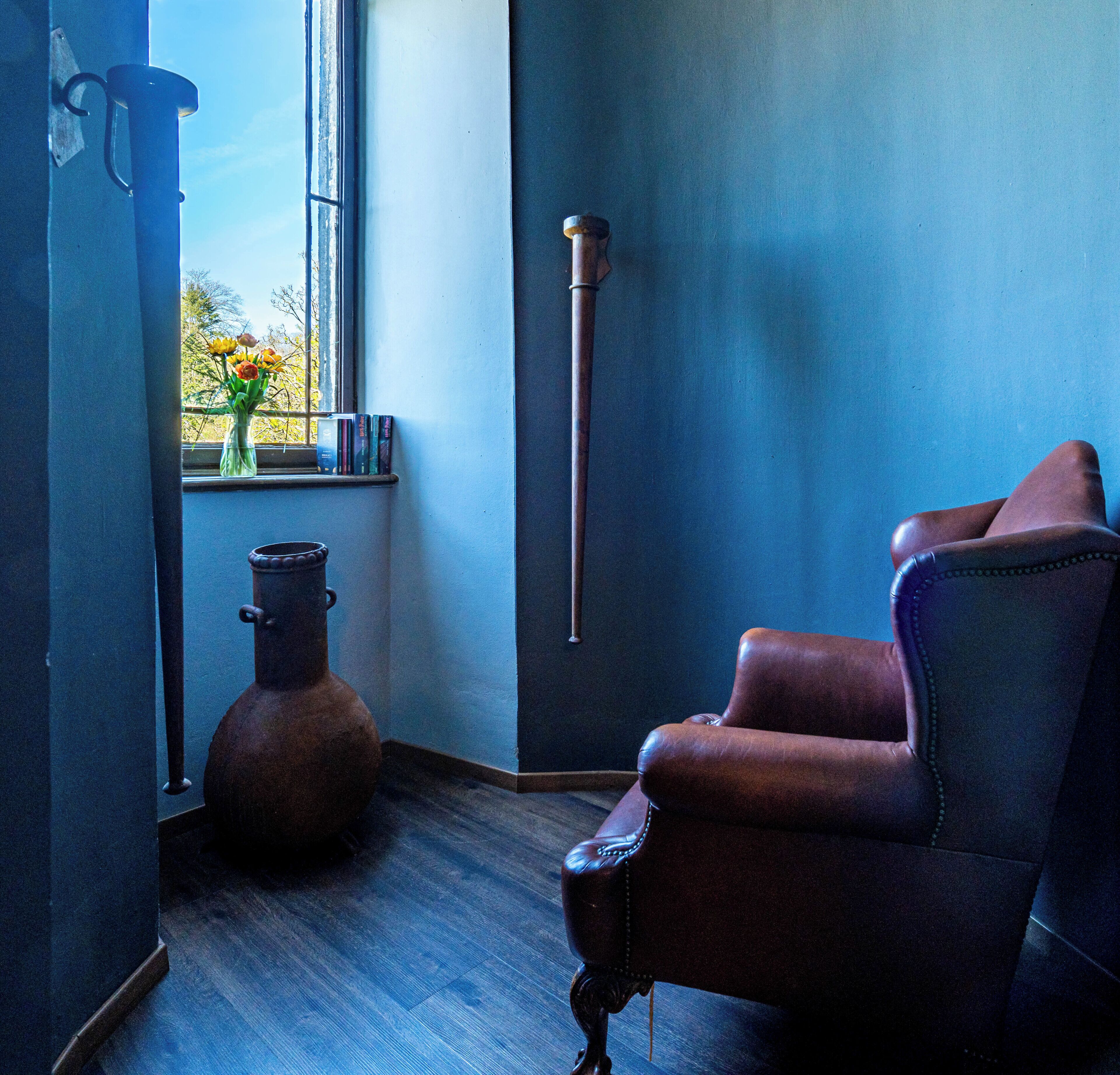 A room with a leather armchair, a large window with a view of trees, a vase with flowers, and a tall, slender object on the wall.