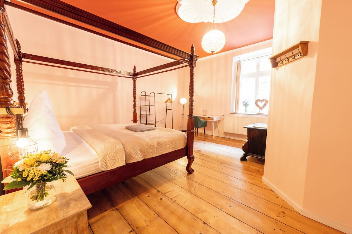 Una cama con dosel de madera, una mesita de noche con un florero, y una ventana con vistas al exterior.