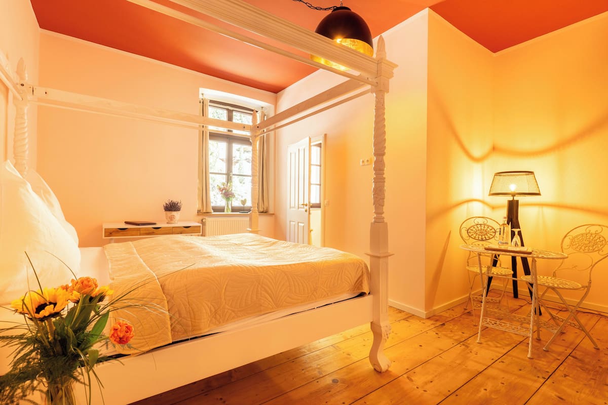 Una cama con dosel, con cabecera blanca, una mesita de noche con lámpara y una pequeña mesa con sillas en una habitación con suelo de madera y techo rojo.