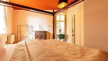 A bedroom with a large bed, a wooden headboard, a desk with a chair, a radiator, and a window with curtains.