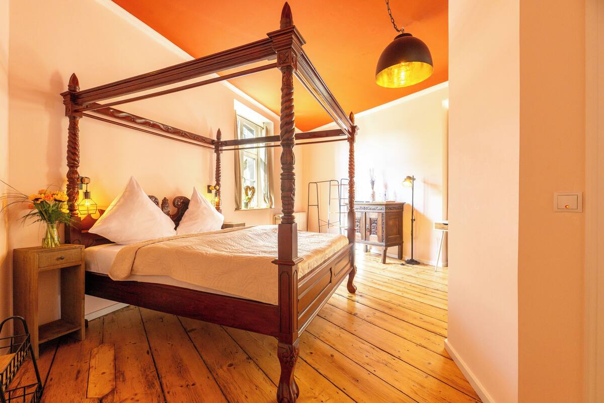 Una cama con dosel de madera y ropa blanca, suelo de madera y lámpara colgante.