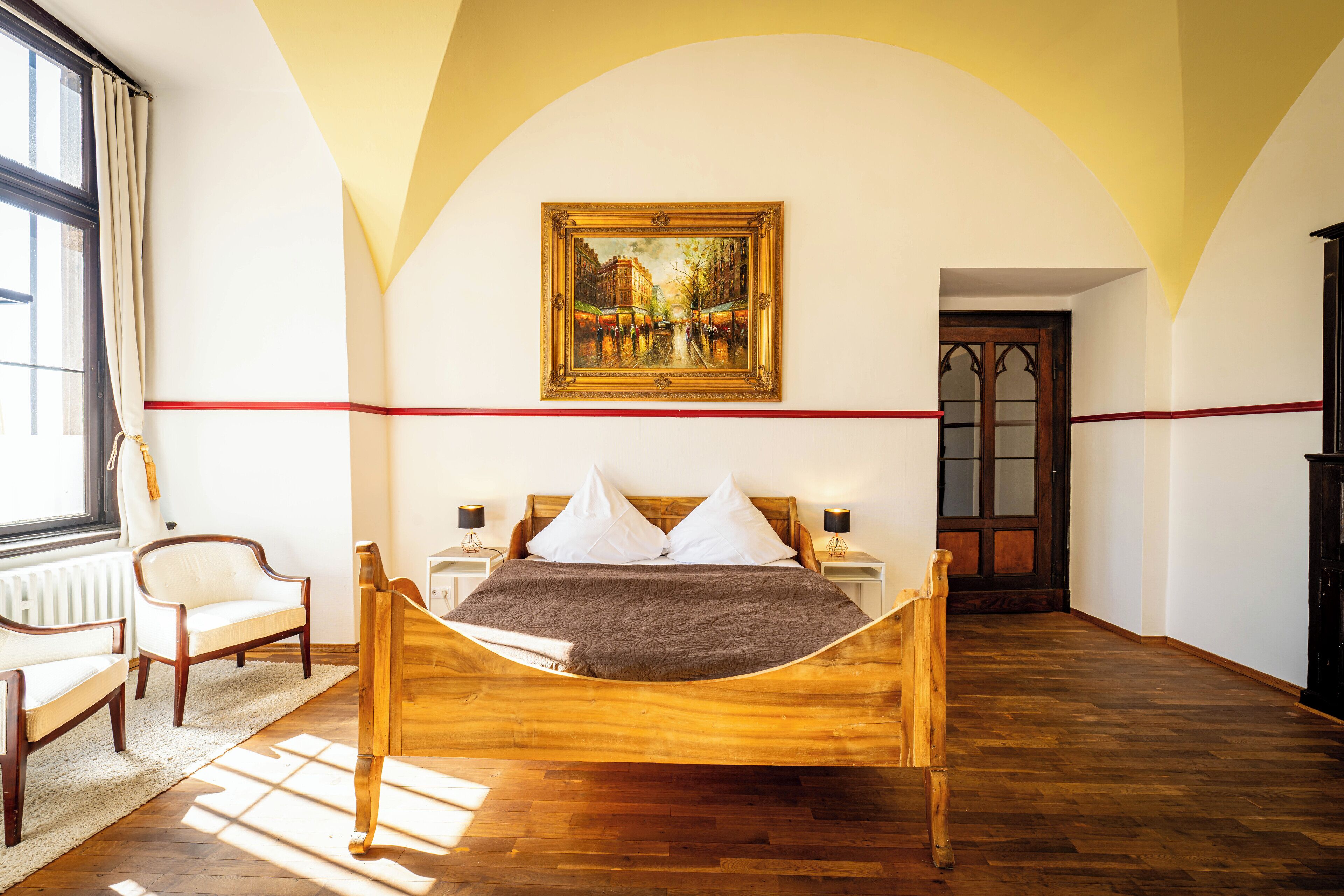 Un dormitorio con cama de madera, almohadas blancas, una franja roja en la pared, un cuadro sobre la cama y una ventana con cortinas.