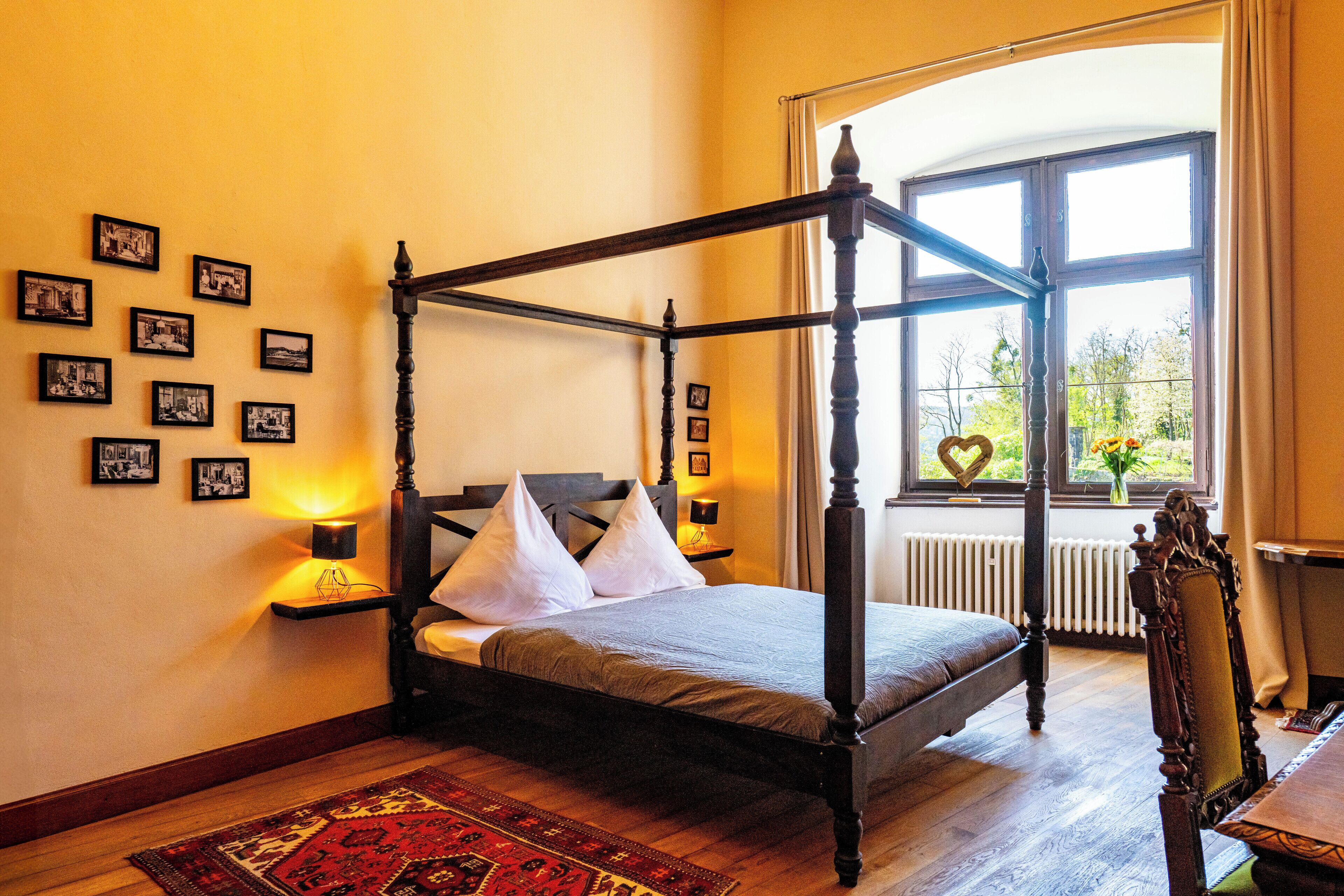 A four-poster bed with white pillows, a red and gold patterned rug, and a series of black and white photographs on the wall.