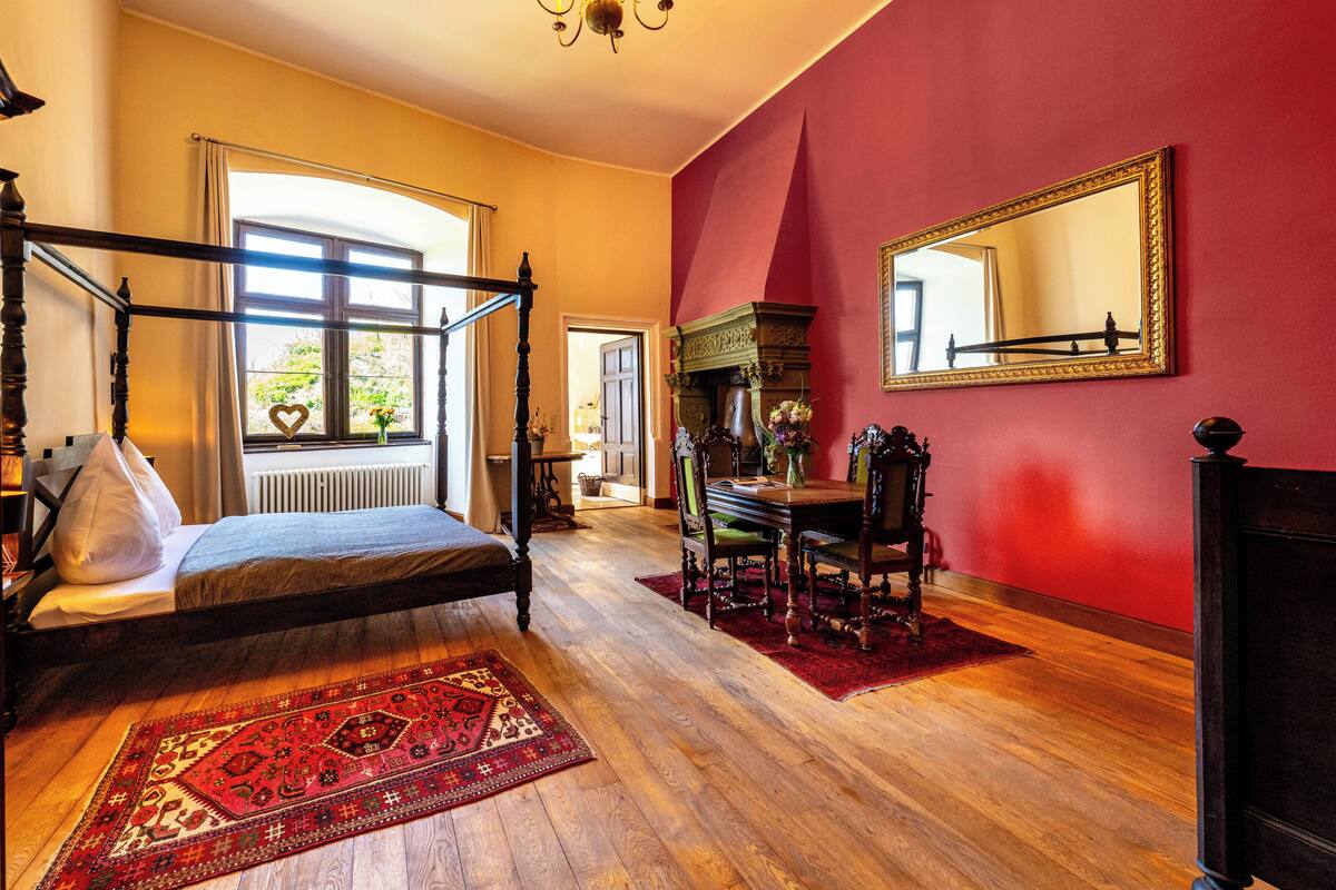 Un dormitorio con cama con dosel, una mesa de comedor con sillas, un candelabro, un espejo grande y una pared roja decorativa.