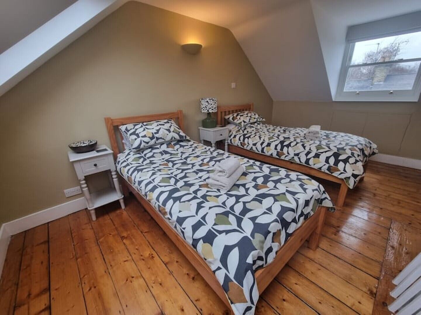Ein Schlafzimmer mit zwei Betten, Holzboden, einem Oberlicht und einem Nachttisch mit Lampe.