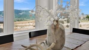 Un vase rustique orné de fleurs blanches, posé sur une table en bois près d’une fenêtre donnant sur un paysage montagneux.