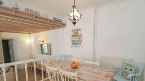 Ein Essbereich mit einem Holztisch, weißen Stühlen und einer Chandelier. Wanddekorationen und ein Blick in einen anderen Raum sind ebenfalls vorhanden.