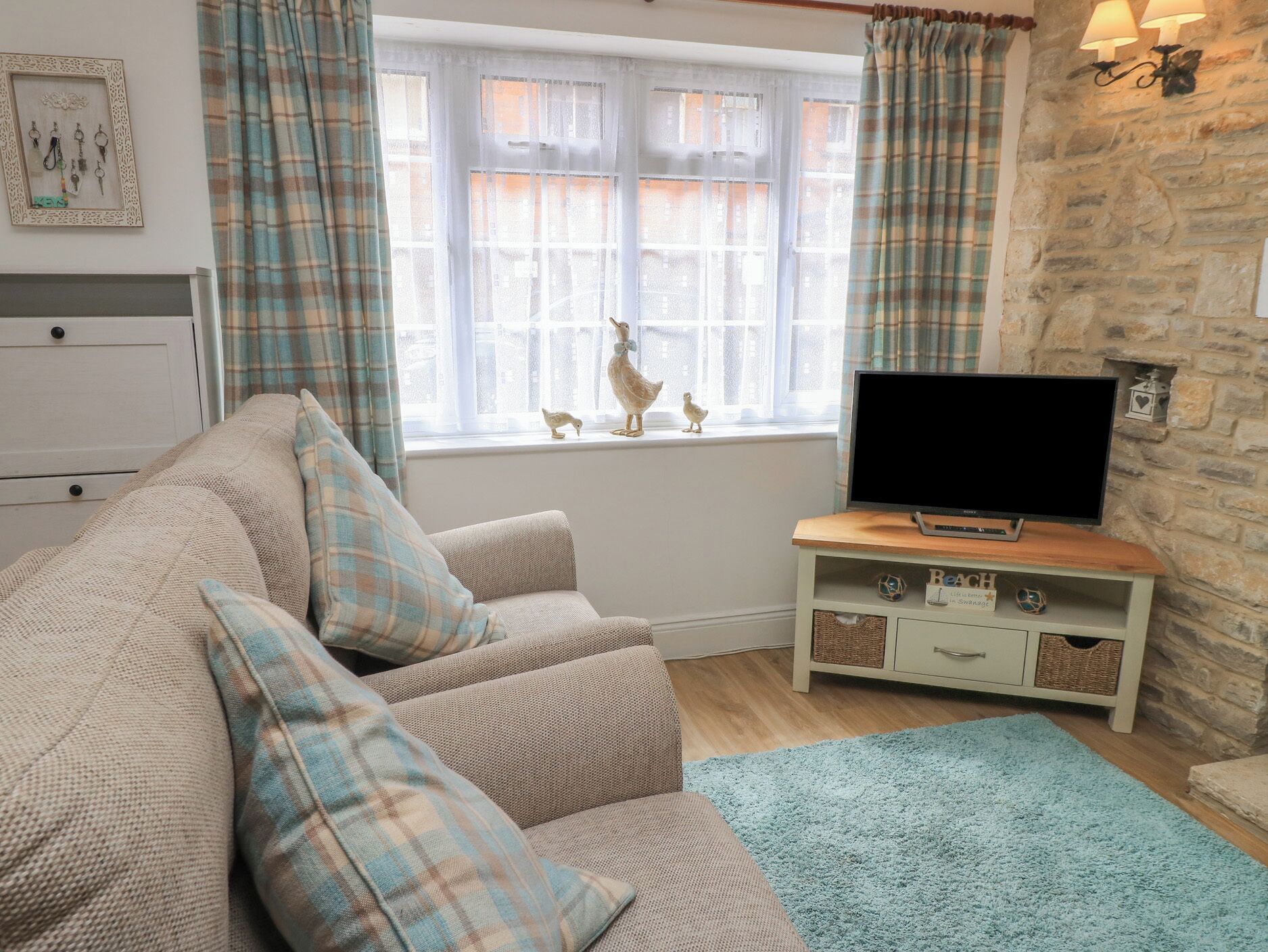 Un salon chaleureux avec un canapé beige, des coussins à carreaux, un téléviseur à écran plat posé sur un meuble en bois, un mur en pierre et une fenêtre avec des rideaux.
