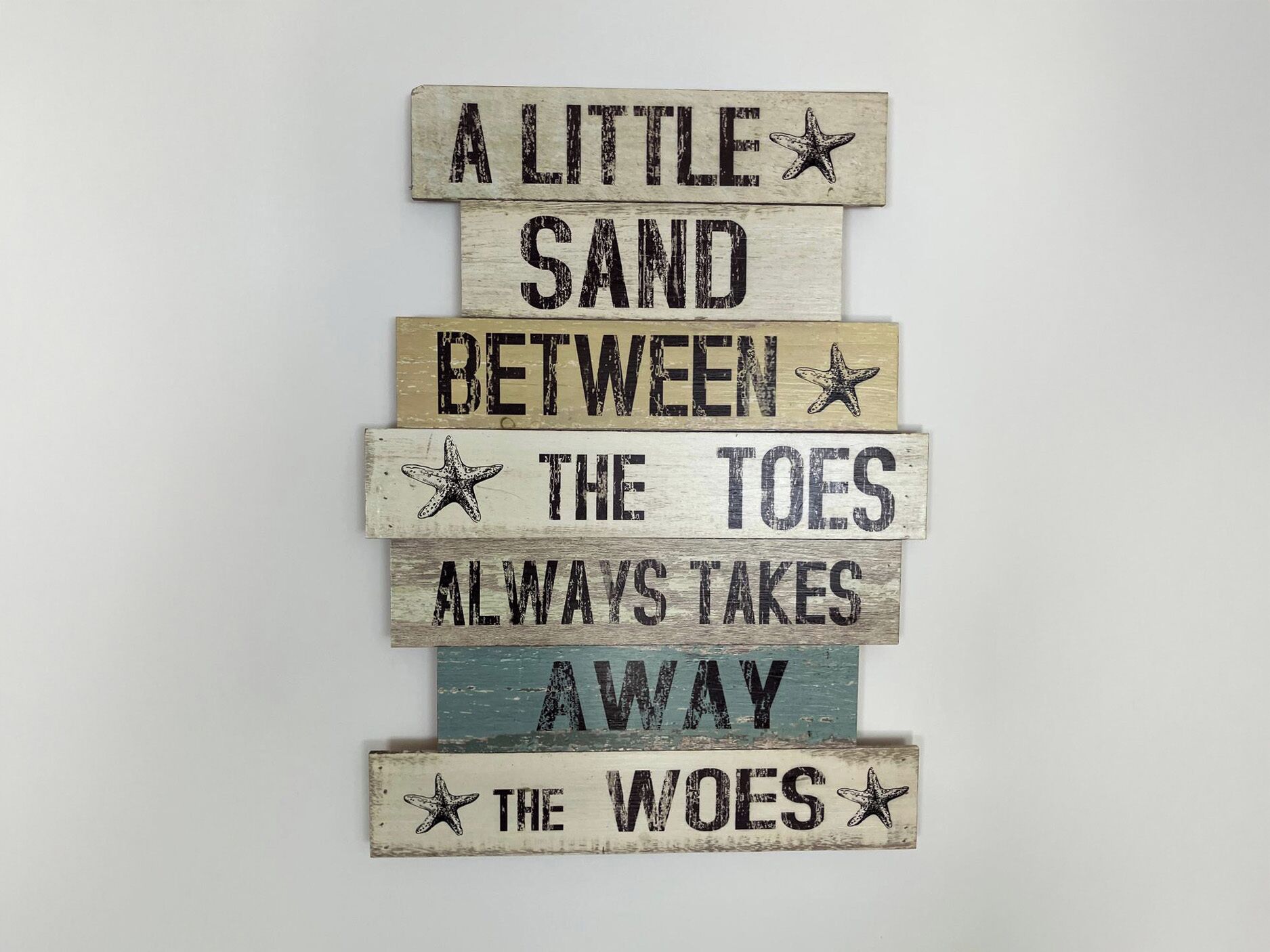 Un letrero de madera con temática náutica, que presenta una estrella de mar y la frase 'Un poco de arena entre los dedos siempre disipa las preocupaciones'.
