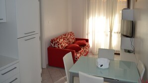 Una habitación pequeña y bien iluminada con un sofá rojo, una mesa de vidrio y un televisor montado en la pared.