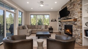 Un salon spacieux avec un canapé d’angle, des fauteuils en cuir, une cheminée et une grande fenêtre donnant sur la forêt.