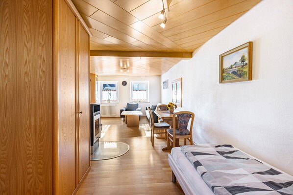 A room with a wooden floor, a bed, a dining table with chairs, a sofa, and a kitchen area with a stove and refrigerator.
