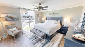A bedroom with a large bed, a chair, a bench, a nightstand, a lamp, a decorative wall piece, and a view of the street through a large window. - Beautiful ST 3025 New Rental! (Destin)