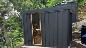 A small, dark-colored cabin with a wooden door, surrounded by a garden with mulch and stepping stones. - Luxury cottage (Port Carling)
