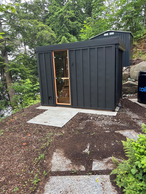 A small, dark-colored cabin with a wooden door, surrounded by a garden with mulch and stepping stones.