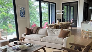 A modern living room with a large sofa, coffee table, and chairs. There is a dining area with a table and chairs visible through large windows. - Luxury cottage (Port Carling)
