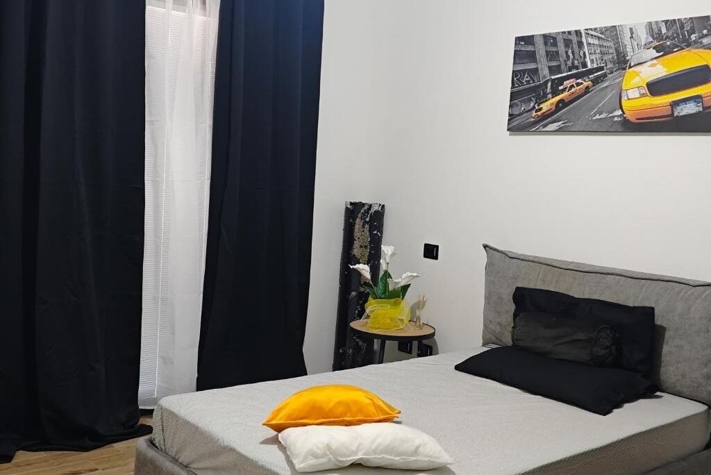 A hotel room with a bed, a small table with a vase of flowers, and a painting of a yellow taxi on the wall.