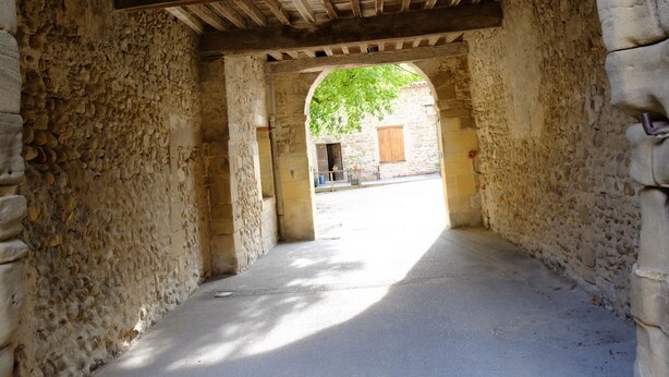 An arched stone passageway with a wooden ceiling, leading to a courtyard with a building in the background.