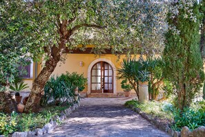 Un camino conduce a un edificio de fachada amarilla, con una entrada en arco y vegetación circundante.