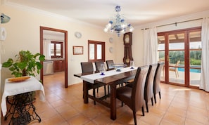 A dining area with a table, chairs, a chandelier, a clock, and a view of a swimming pool. - Country House 'Tolo' with Sea View, Private Pool and Wi-Fi (Son Servera)