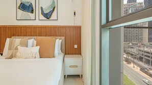 Ein modernes Hotelzimmer mit einem großen Bett, einem Nachttisch und Blick auf die Stadt durch große Fenster.
