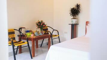 A room with a bed, a wooden table with chairs, a potted plant, and a wall-mounted shelf.