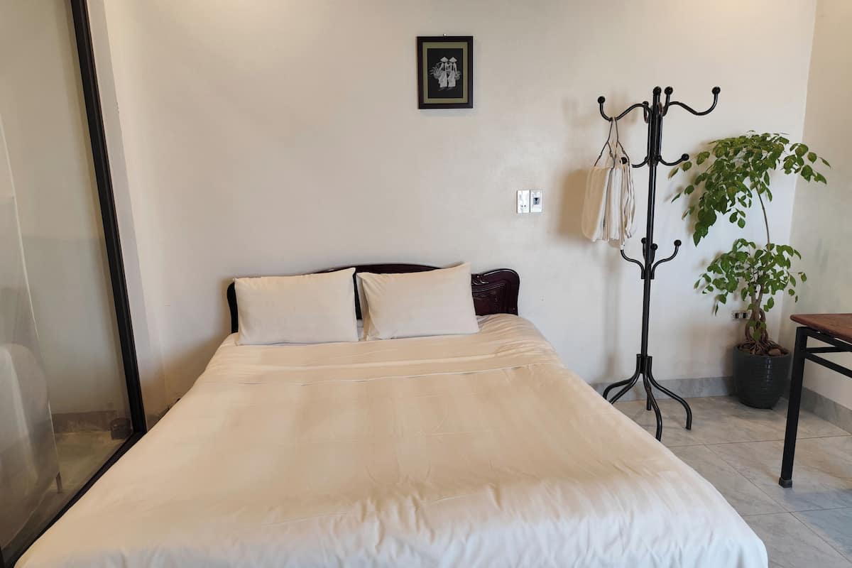 A neatly made bed with a headboard, a potted plant, and a coat rack in a room with a plain wall and a framed picture.