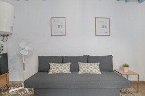 Une chambre avec un canapé gris, deux coussins à motifs, un ventilateur sur pied, une petite table avec une plante et deux tableaux encadrés accrochés au mur.