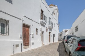 Une ruelle étroite pavée, bordée de bâtiments blancs, avec des balcons et une rangée de voitures garées.