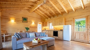 A wooden cabin interior with a sofa, dining table, kitchen area, and large windows.