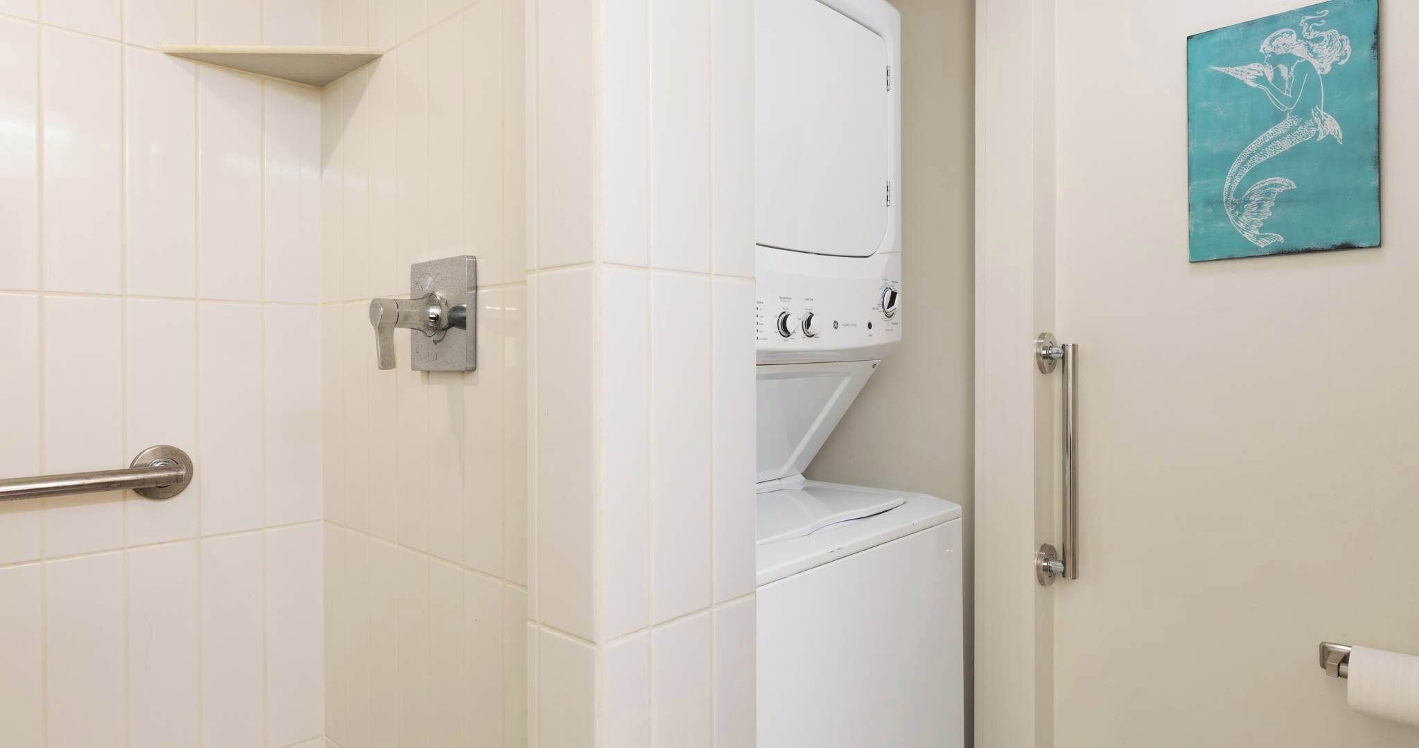 Une salle de bain avec des carreaux blancs, une barre d’appui, un porte-serviettes et un distributeur de serviettes fixé au mur.