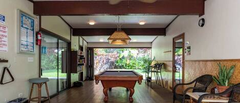 A room with a pool table, bookshelves, and a view of a garden.