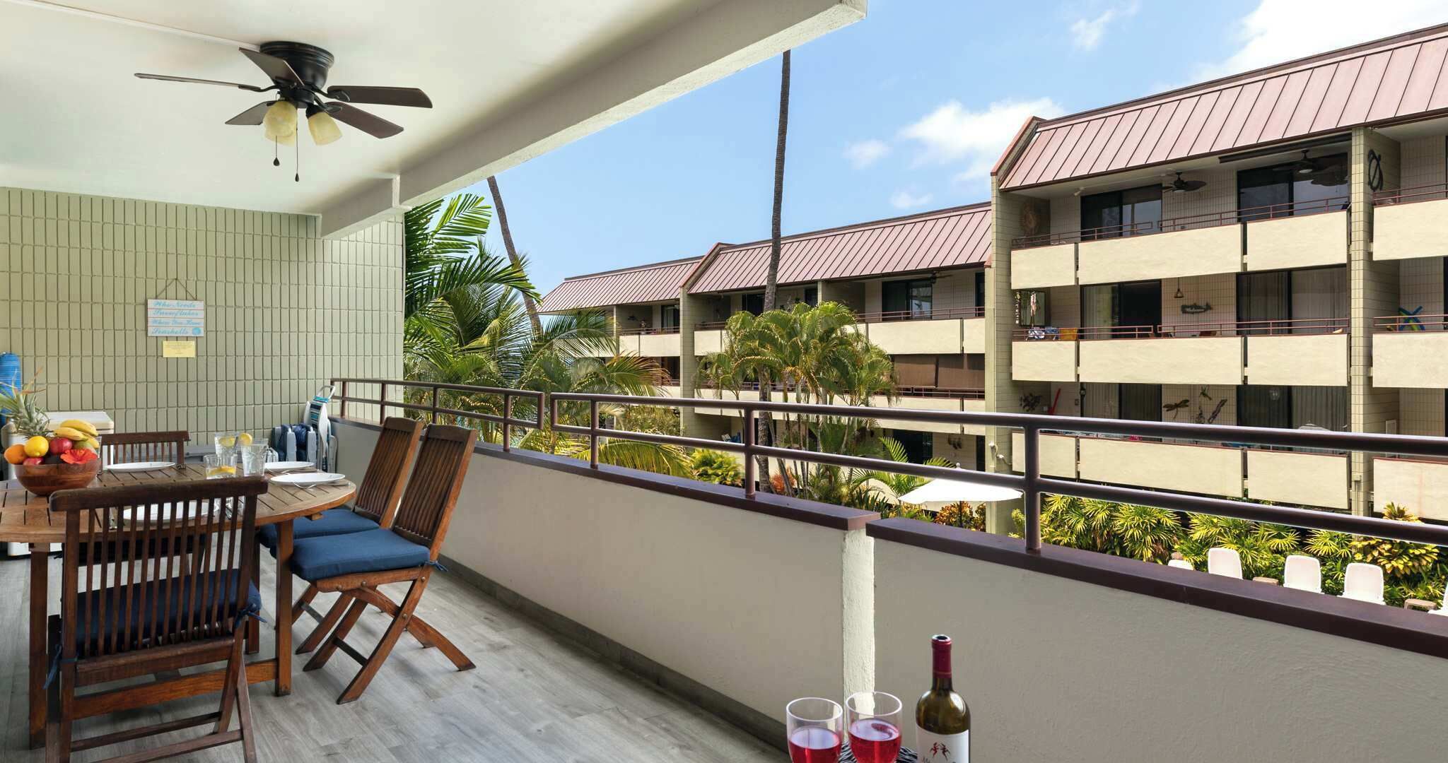 A balcony with a wooden table, chairs, and a fruit bowl. There are two wine glasses and a bottle on the table. The balcony overlooks a building with multiple balconies and palm trees.