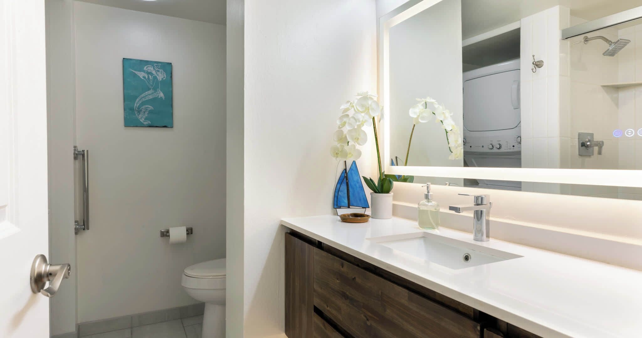 Un baño moderno con un lavamanos blanco, un espejo y un inodoro. Hay una obra de arte azul en la pared y orquídeas blancas en la encimera.