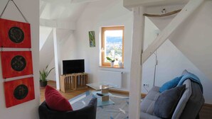 A living room with a TV on a wooden stand, a couch, a red cushion, a glass coffee table, and a view of a city through a window. - Apartment 'Loft Straussi' with Shared Garden and Wi-Fi (Tuttlingen)
