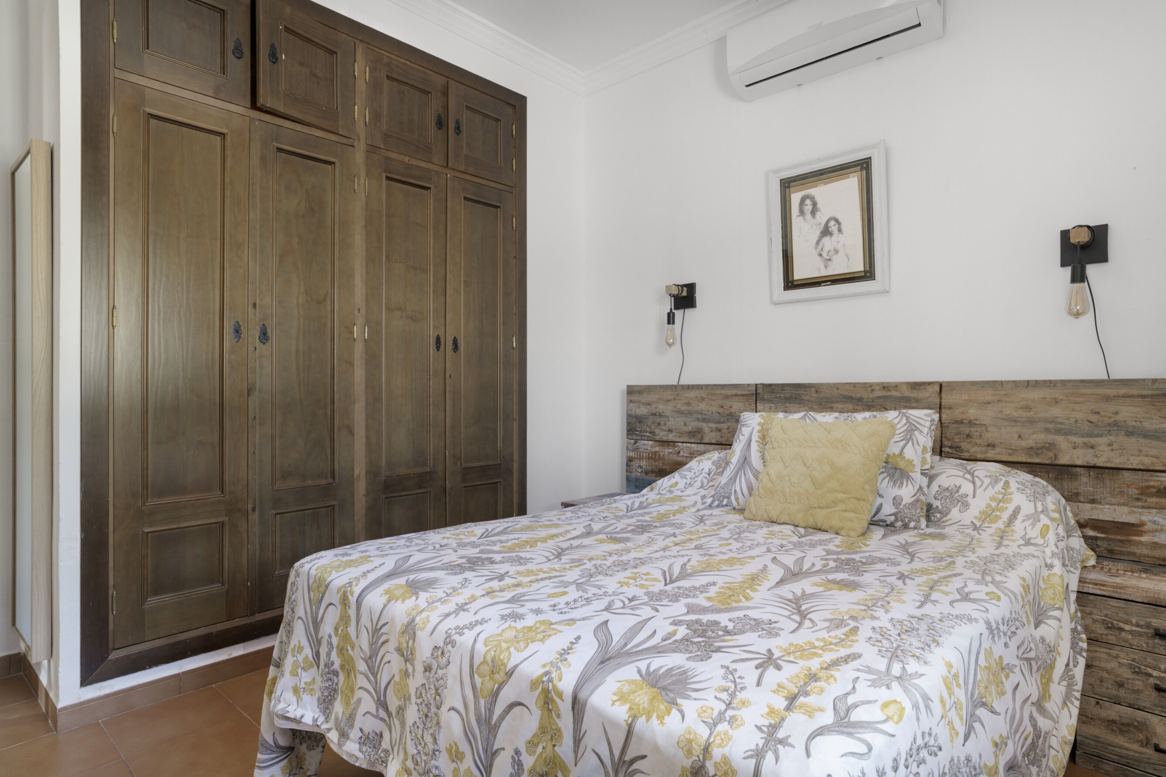 Een slaapkamer met een bed, een houten garderobe, een wandairconditioner en een lijst met een afbeelding.