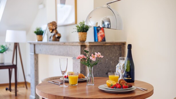 Ein runder Holztisch mit Frühstücksangeboten, darunter Croissants, Obst, Saft und eine Weinflasche.