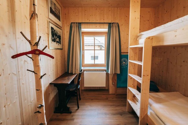 A wooden room with a table, chairs, a bunk bed, a window with curtains, and a coat rack.