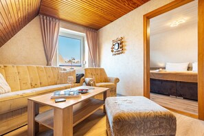 A cozy room with a wooden ceiling, a sofa, two armchairs, a small table with glasses and a remote control, and a bed with a headboard.