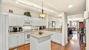 A modern kitchen with white cabinets, a central island, and a dining area with a red lamp. - Kings Grant 203- 2BR Ocean Drive- Pool, Hot Tub (North Myrtle Beach)