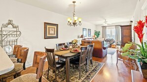 A dining area with a wooden table, chairs, and a chandelier. There is a sofa and a plant in the background. - Kings Grant 203- 2BR Ocean Drive- Pool, Hot Tub (North Myrtle Beach)