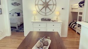 Una habitación con una mesa de comedor de madera, un tocador blanco, una cama con cabecera azul y un adorno de brújula colgado en la pared.