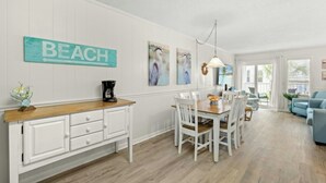 A dining area with a wooden table and white chairs, a white cabinet with drawers, and a wall-mounted sign that reads "BEACH". - Finestere G1- 2BR in Cherry Grove- Pool (North Myrtle Beach)