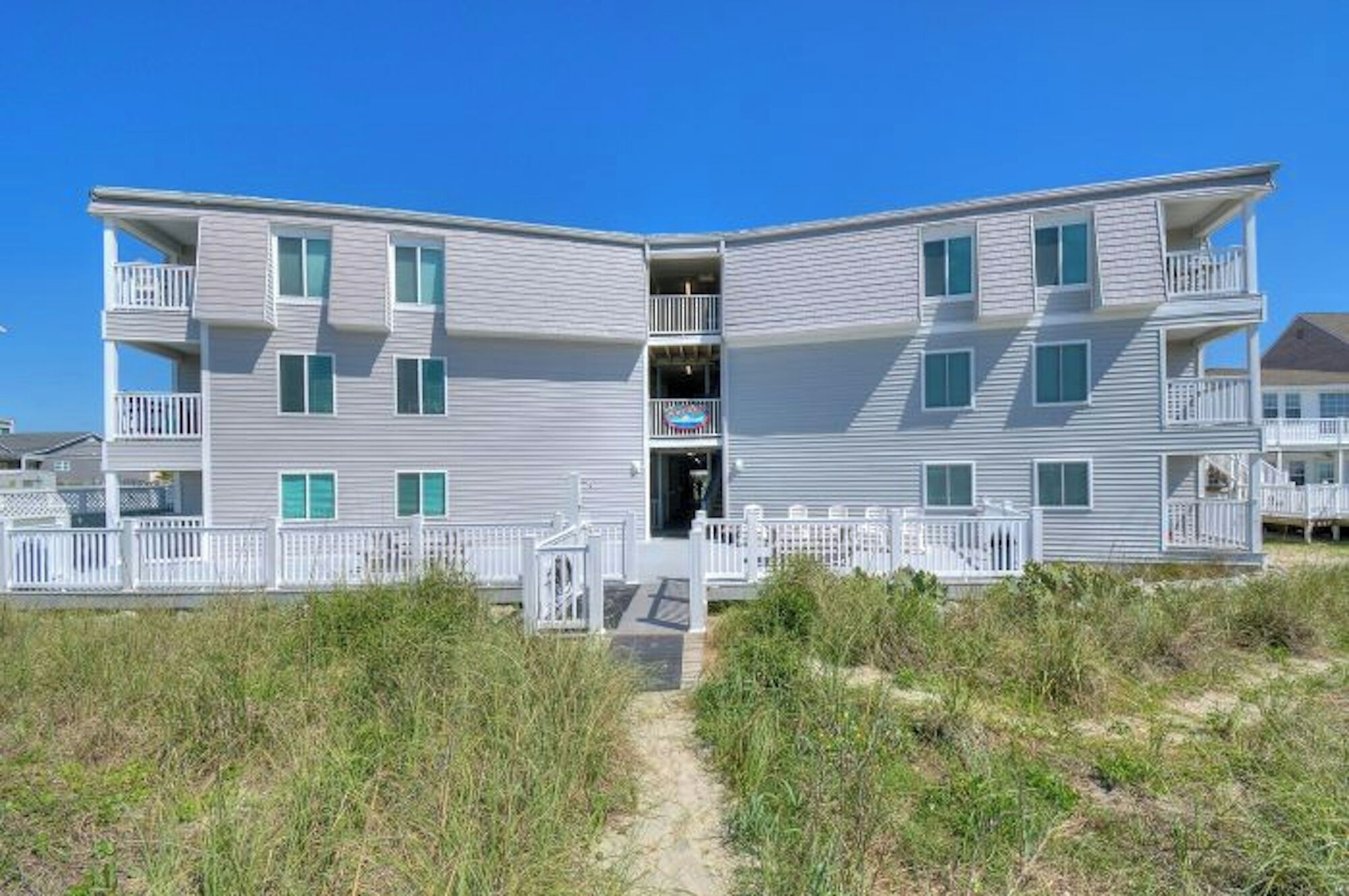 A two-story beachfront building with balconies, a central entrance, and a sandy path leading to the entrance.