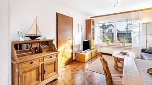 Ein Wohnzimmer mit Holzschrank, Fernseher, Esstisch und Fenster mit Blick auf Gebäude.