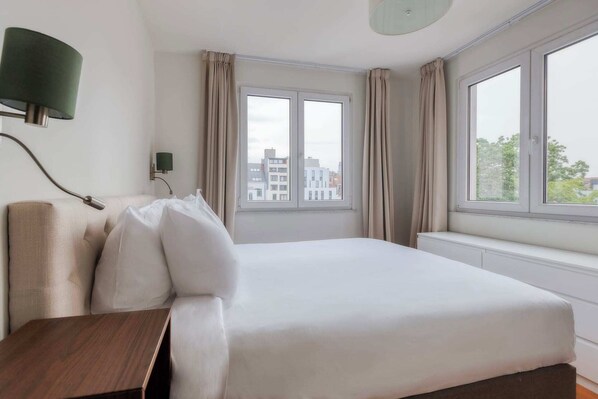 A hotel room with a bed, a nightstand, a lamp, and a view of buildings through the windows.