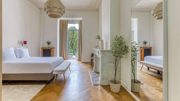 A spacious bedroom with a large bed, a small table, a fireplace, and a balcony with a view.