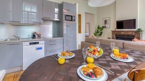 A modern kitchen with grey cabinets, a built-in microwave, and a dishwasher. There is a dining table set with plates of breakfast, including croissants, fruit, and juice. A sofa and a TV are visible in the living area.