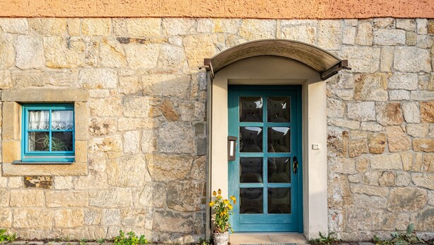 Un bâtiment en pierre avec une porte et une fenêtre bleues, un pot de fleurs et une grille métallique à l’entrée.
